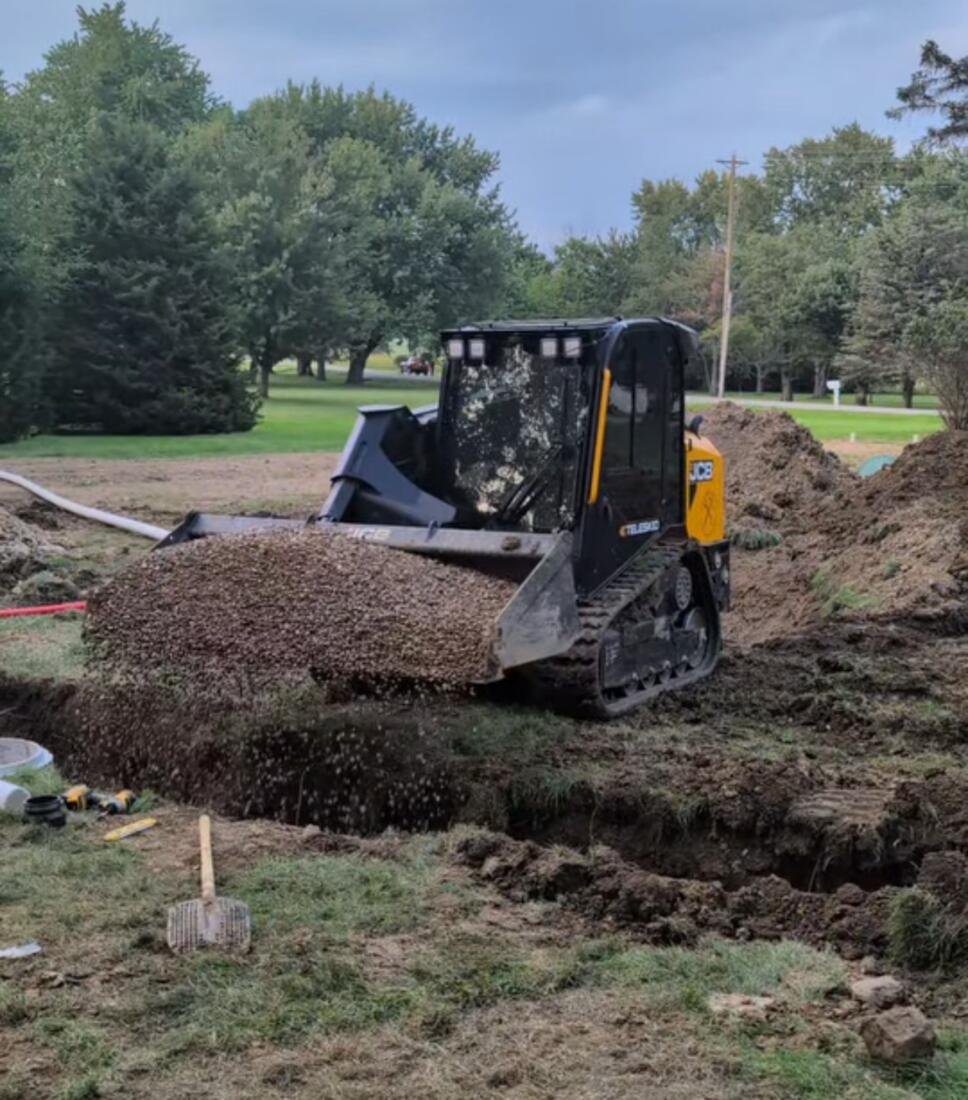 Skid steer spreading stone into a drainage trench next to a house